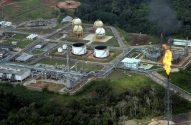 Complexo industrial com tanques e tubulações, cercado por floresta, com duas torres de flare queimando gás.