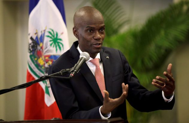 Homem de terno fala ao microfone, gesticulando. Ao fundo, bandeira do Haiti e folhagem verde.
