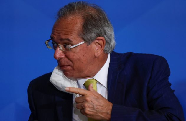 Homem de terno e óculos, com máscara branca abaixada, aponta para ela.