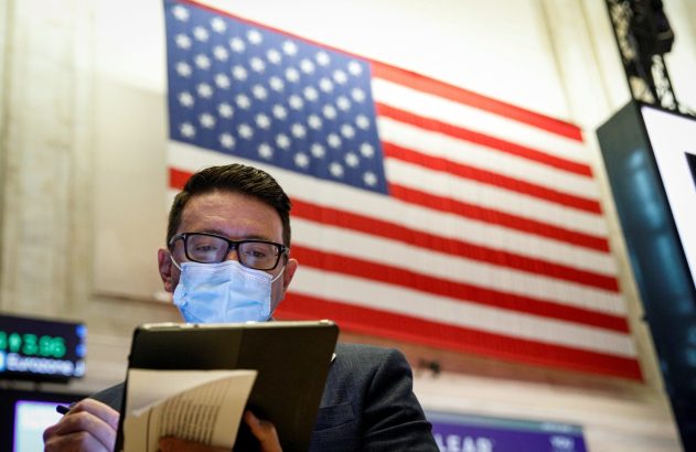 Homem de óculos e máscara escreve em tablet, com bandeira dos EUA e tela digital ao fundo.