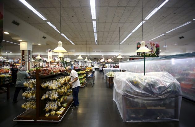 Supermercado com clientes comprando. Prateleiras de frutas e verduras, algumas cobertas por plástico. Decoração de Páscoa ao fundo.