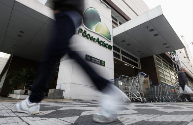 Pessoa desfocada andando em frente à entrada do Pão de Açúcar, com carrinhos de compras estacionados.