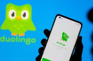 Mão segura smartphone com app Duolingo na tela. Logo Duolingo e nome da marca aparecem grandes no fundo azul.