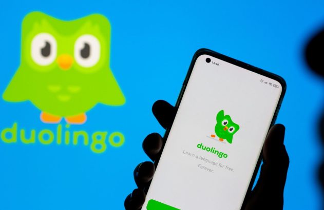 Mão segura smartphone com app Duolingo na tela. Logo Duolingo e nome da marca aparecem grandes no fundo azul.