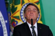 Jair Bolsonaro, de terno, fala ao microfone. Bandeiras do Brasil e azul ao fundo.