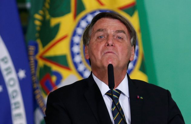 Jair Bolsonaro, de terno, fala ao microfone. Bandeiras do Brasil e azul ao fundo.
