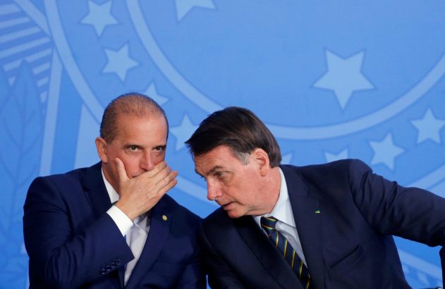 Dois homens de terno. Um fala ao ouvido do outro, que cobre a boca com a mão. Fundo azul com estrelas.