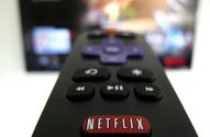 Controle remoto preto com o botão Netflix em primeiro plano, e uma TV com tela desfocada ao fundo.
