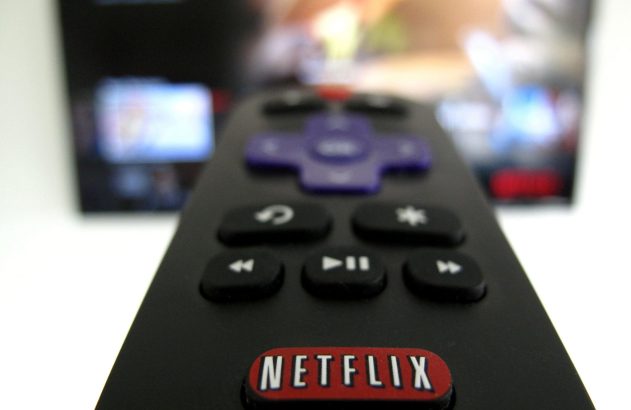 Controle remoto preto com o botão Netflix em primeiro plano, e uma TV com tela desfocada ao fundo.