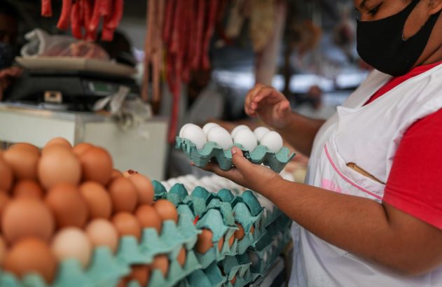 Mulher com máscara e avental organiza ovos brancos em cartela. Ovos marrons empilhados e carnes ao fundo.