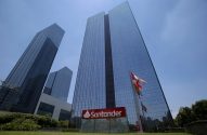 Edifício Santander de vidro, com logo vermelho, bandeiras e céu azul.