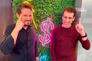Dois homens sorrindo. Um aponta para a têmpora, o outro para cima. Fundo verde com sinal de dólar em neon rosa.