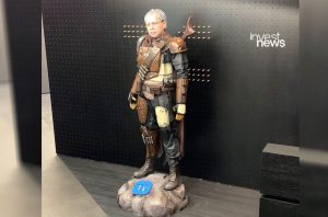 Estátua de homem de óculos e cabelo grisalho com armadura Mandaloriana e rifle. Logo 'invest news' ao fundo.