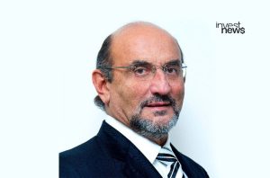 Homem de óculos e barba, usando terno e gravata listrada, olhando para a câmera. Fundo branco.