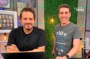 Dois apresentadores do Invest News em estúdio, um com laptop e outro de pé, ambos sorrindo para a câmera.