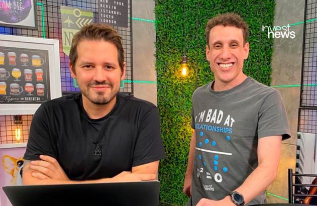 Dois apresentadores do Invest News em estúdio, um com laptop e outro de pé, ambos sorrindo para a câmera.