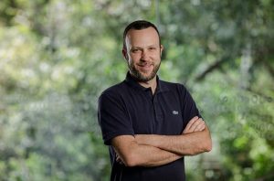 Homem barbudo sorrindo de polo azul escuro e braços cruzados, em frente a fundo verde desfocado.