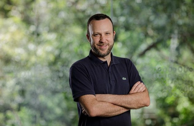 Homem barbudo sorrindo de polo azul escuro e braços cruzados, em frente a fundo verde desfocado.