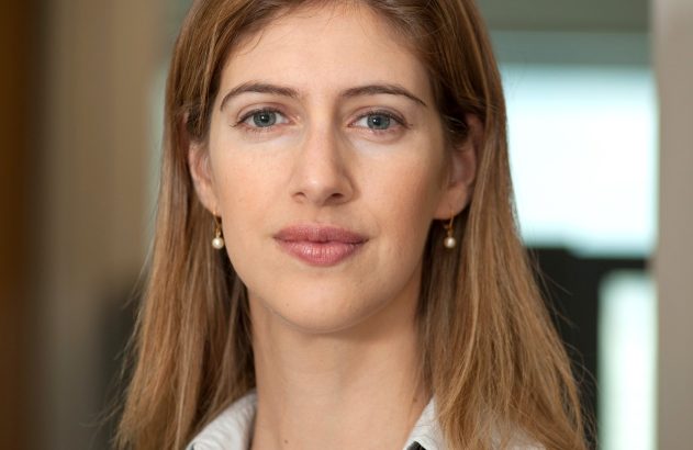Retrato frontal de mulher com cabelo loiro, olhos azuis, terno preto e brincos de pérola.