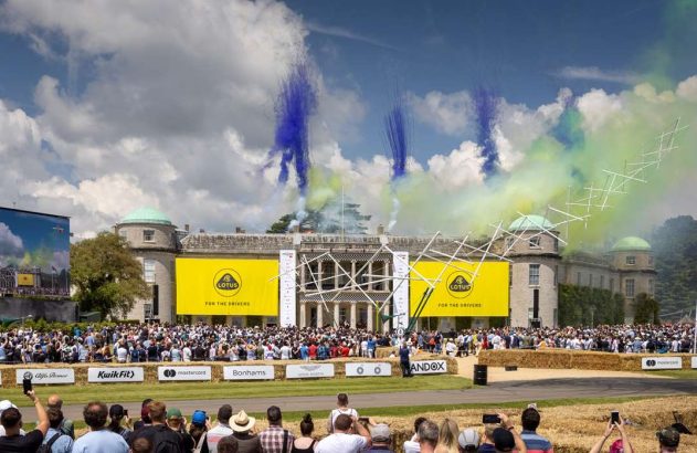 Multidão em evento ao ar livre assiste a exibição com fumaça colorida e estrutura metálica Lotus sobre mansão. Banners Lotus.