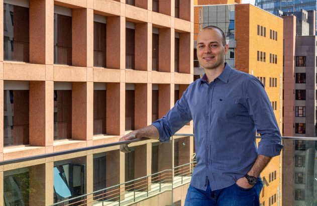 Homem careca sorrindo, camisa azul e jeans, apoiado em corrimão. Edifícios modernos ao fundo.
