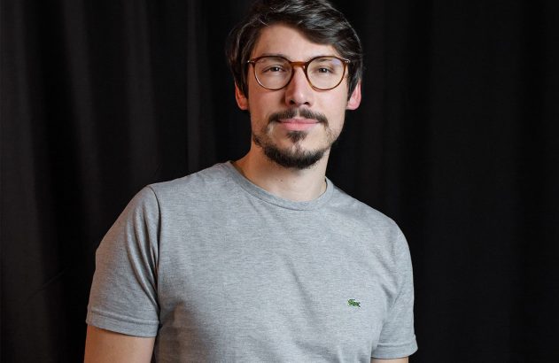 Homem com óculos e barba em camiseta cinza sorri. Fundo preto.