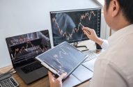 Homem analisando gráficos de ações em computador e monitor, apontando com caneta e segurando prancheta.