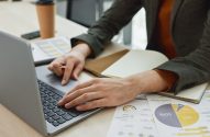Mãos em laptop, trabalhando com documentos e gráficos de análise de dados em mesa de escritório.