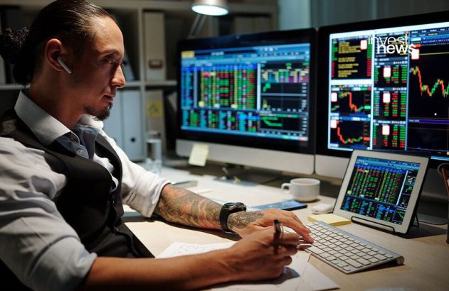 Homem com fone, focado em monitores de trading com gráficos e dados, fazendo anotações.