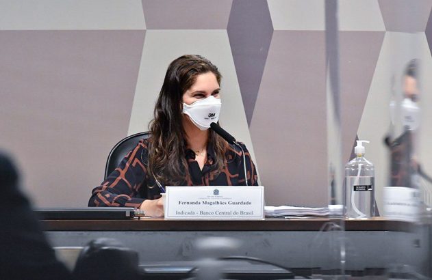 Fernanda Magalhães Guardado, indicada ao Banco Central do Brasil, sentada à mesa, de máscara, escrevendo e falando.