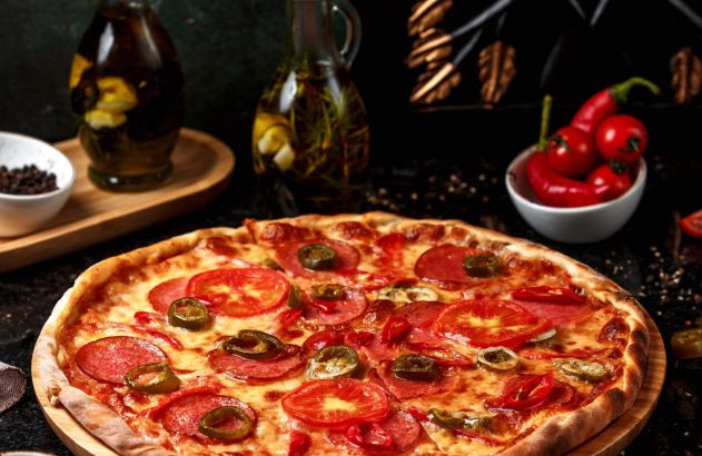 Pizza com salame, tomate e jalapeño em tábua de madeira. Garrafas de azeite e tigelas de temperos ao fundo.