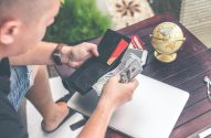 Homem contando dinheiro da carteira com notas de dólar, laptop e globo em mesa de madeira.