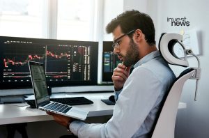 Homem com óculos e camisa, analisando gráficos de trade em laptop e monitores no escritório.