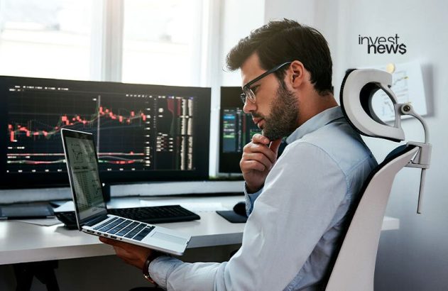 Homem com óculos e camisa, analisando gráficos de trade em laptop e monitores no escritório.