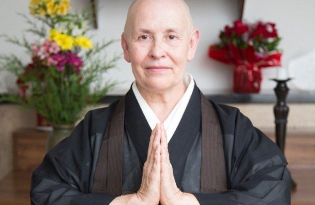 Pessoa careca em vestes escuras e gola branca, mãos juntas em oração/saudação. Flores ao fundo.