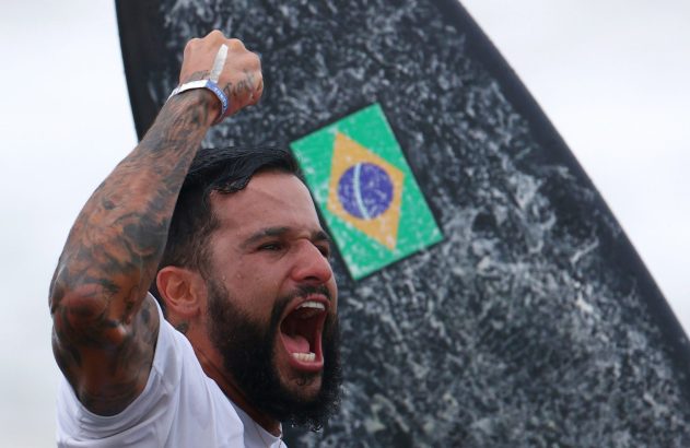 Surfista brasileiro comemorando com punho erguido e boca aberta. Bandeira do Brasil em sua prancha.