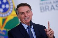Bolsonaro, com sorriso largo, fala ao microfone e levanta o dedo. Bandeira do Brasil ao fundo.