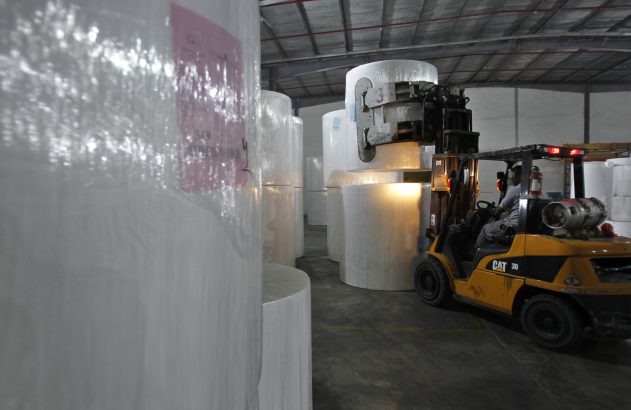 Empilhadeira amarela CAT com operador movimenta rolo branco gigante em armazém. Inúmeras outras bobinas preenchem o galpão.