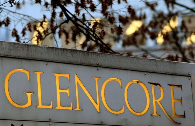 Placa cinza com o nome 'GLENCORE' em letras douradas. No fundo, galhos de árvores e luzes embaçadas.