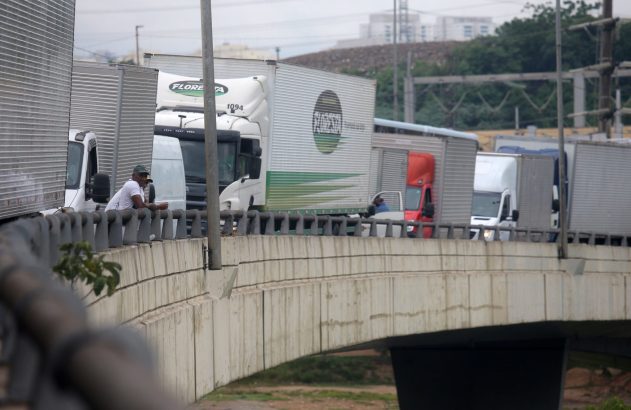 Longa fila de caminhões e vans parados em congestionamento sobre uma ponte de concreto. Um motorista observa.