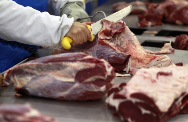 Mão com luva e faca cortando carne crua em uma bancada.