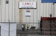 Fábrica JBS Greeley Beef Plant. Um letreiro grande, alambrado e três pessoas, sendo uma em uniforme de trabalho, na área de entrada.