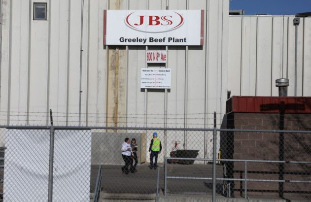 Prédio cinza da JBS Greeley Beef Plant com letreiro e pessoas na entrada. Cerca de arame farpado em primeiro plano.