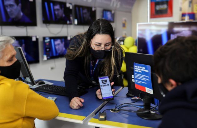 Atendente em loja de eletrônicos mostra celular a cliente. Todos usam máscaras; TVs ao fundo.