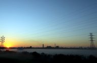 Amanhecer sobre paisagem enevoada com torres e linhas de energia. Ao fundo, silhueta industrial.