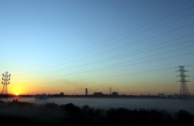 Amanhecer sobre paisagem enevoada com torres e linhas de energia. Ao fundo, silhueta industrial.