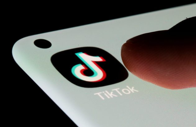Um dedo toca a tela de um smartphone exibindo o ícone e nome do app TikTok, com furo da câmera visível.