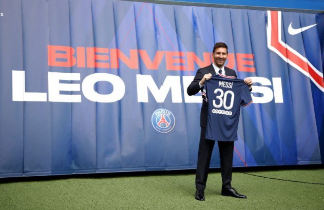 Messi sorri, segurando camisa 30 do PSG, com banner "BIENVENUE LEO MESSI" atrás.