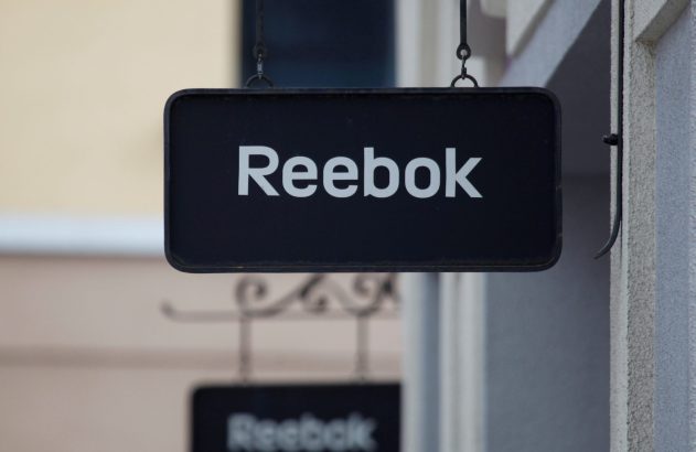 Letreiro preto da Reebok com letras brancas pendurado. Um segundo letreiro da marca, desfocado, é visível abaixo.