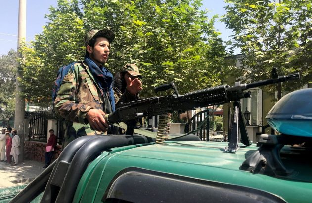 Homem de uniforme militar com metralhadora em veículo verde. Outro atrás. Pessoas em calçada. Dia ensolarado.